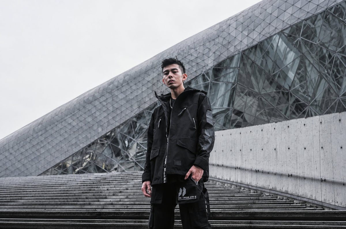A man wearing a black tactical-style jacket and matching black pants stands on outdoor concrete stairs in front of a modern geometric glass building.